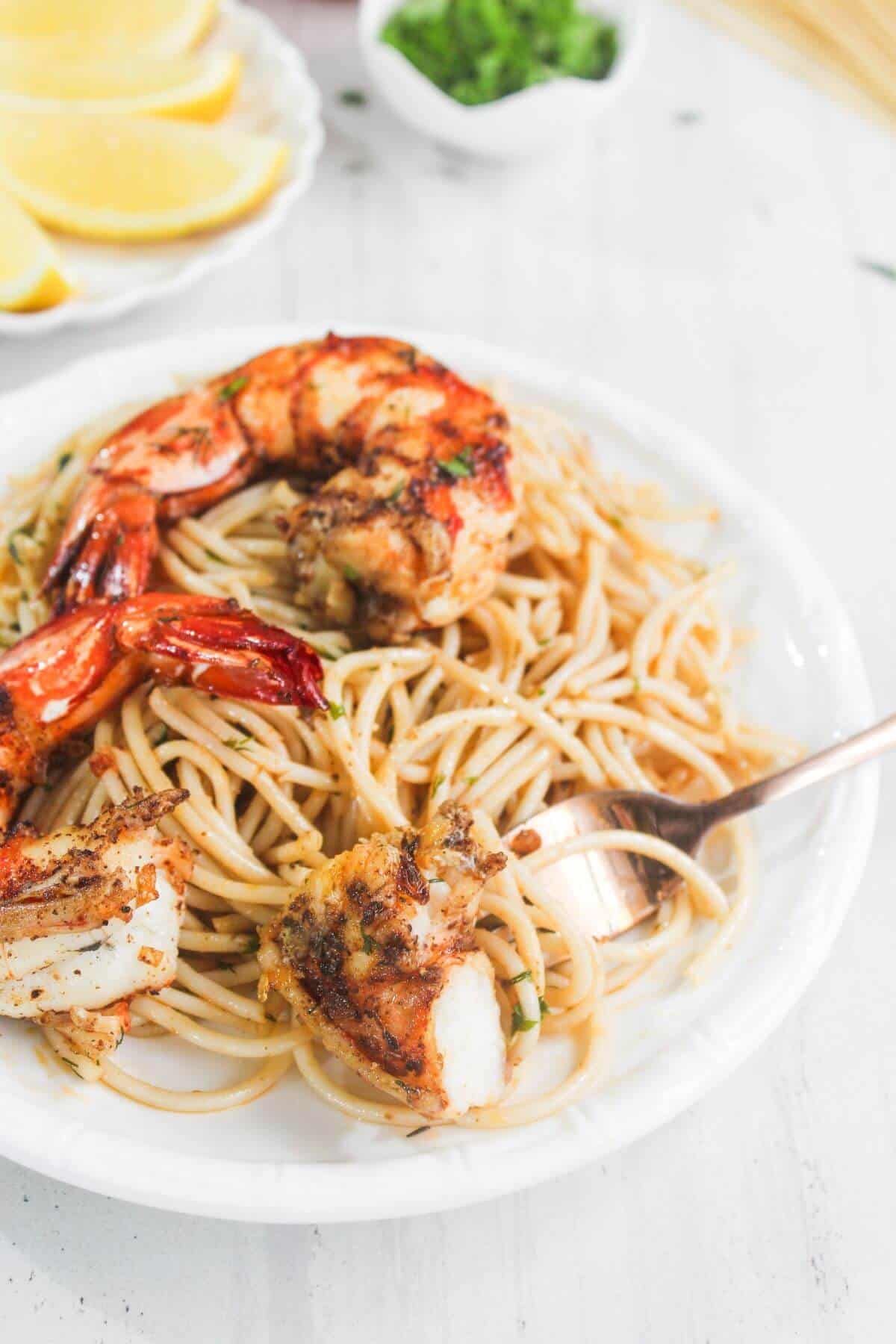 A white plate of spaghetti topped with grilled shrimp, with a fork resting on the side. Lemon slices and chopped herbs are visible in the background.