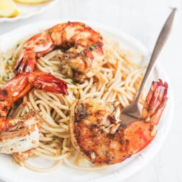 Grilled shrimp served on a bed of spaghetti with herbs, presented on a white plate with a fork.
