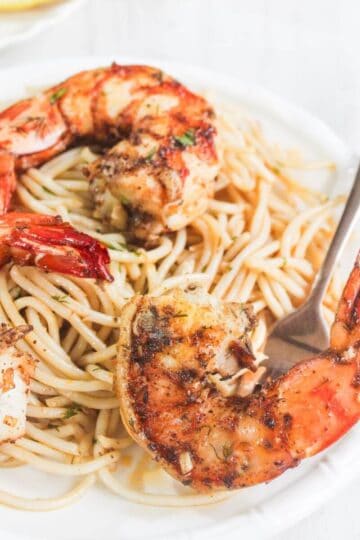Grilled shrimp served on a bed of spaghetti with herbs, presented on a white plate with a fork.