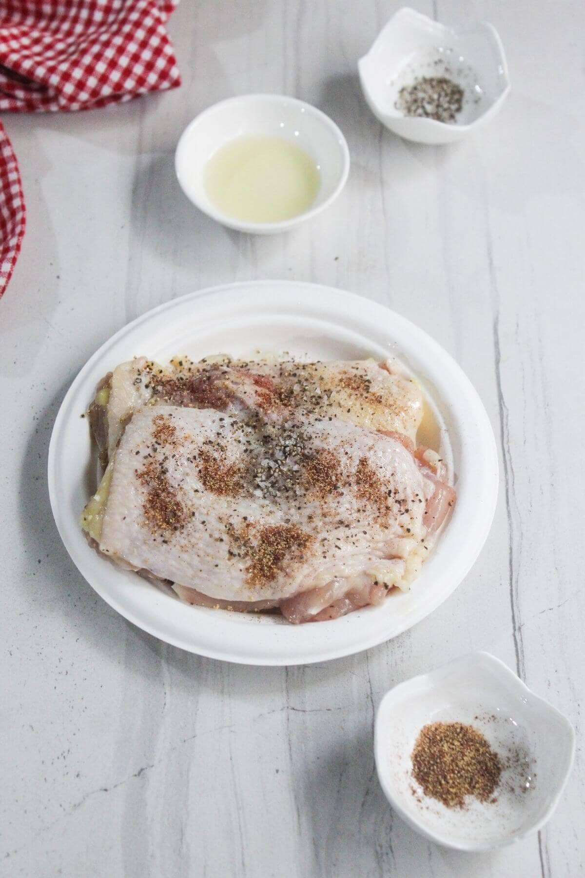 Raw chicken pieces on a white plate sprinkled with spices, surrounded by small bowls of seasoning and a bowl of liquid on a light surface.