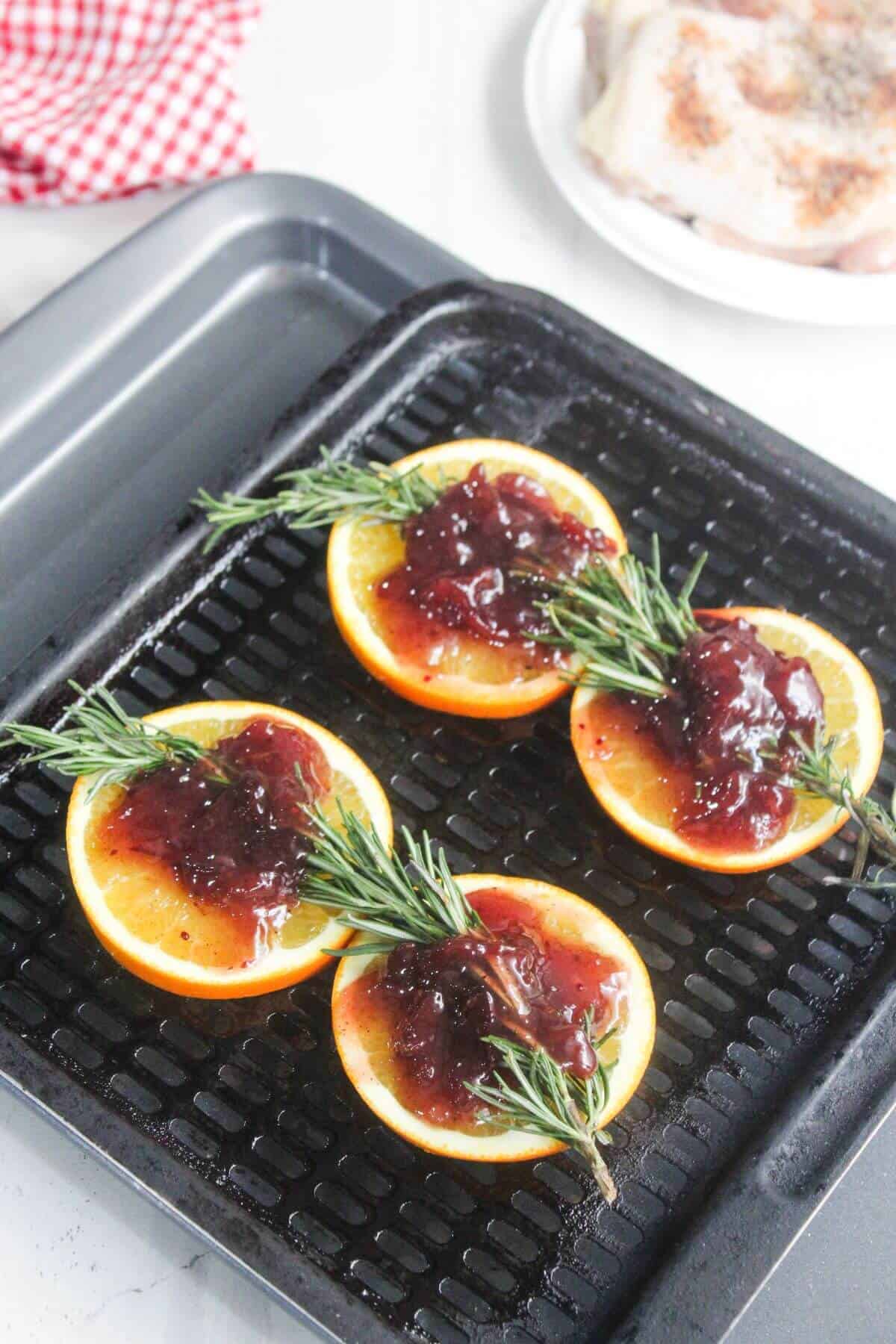 Four orange slices topped with a dark red sauce and sprigs of rosemary are arranged on a grill pan. A plate of raw seasoned chicken is visible in the background.