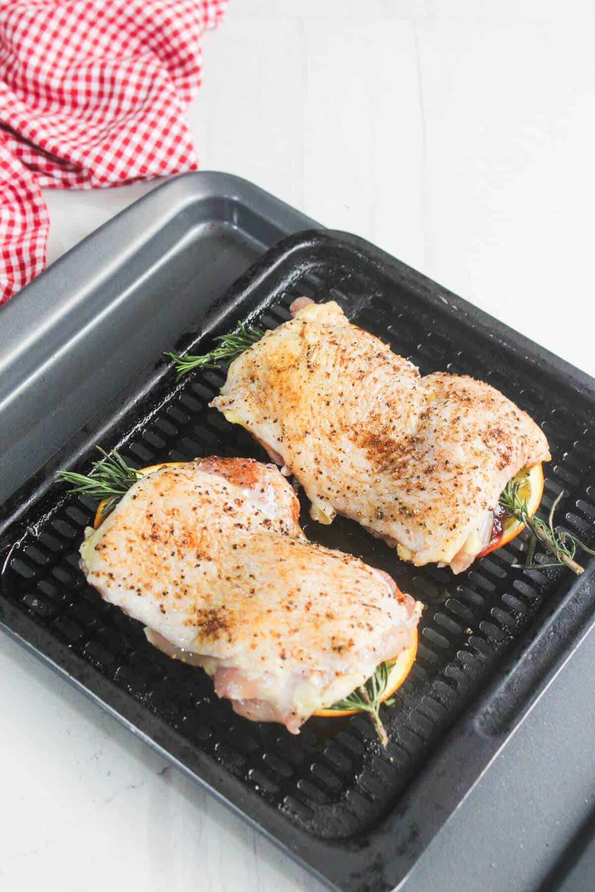 Two raw, seasoned chicken breasts sit on a black roasting rack, stuffed with herbs and lemon slices, ready for cooking. A red and white checkered towel is nearby.