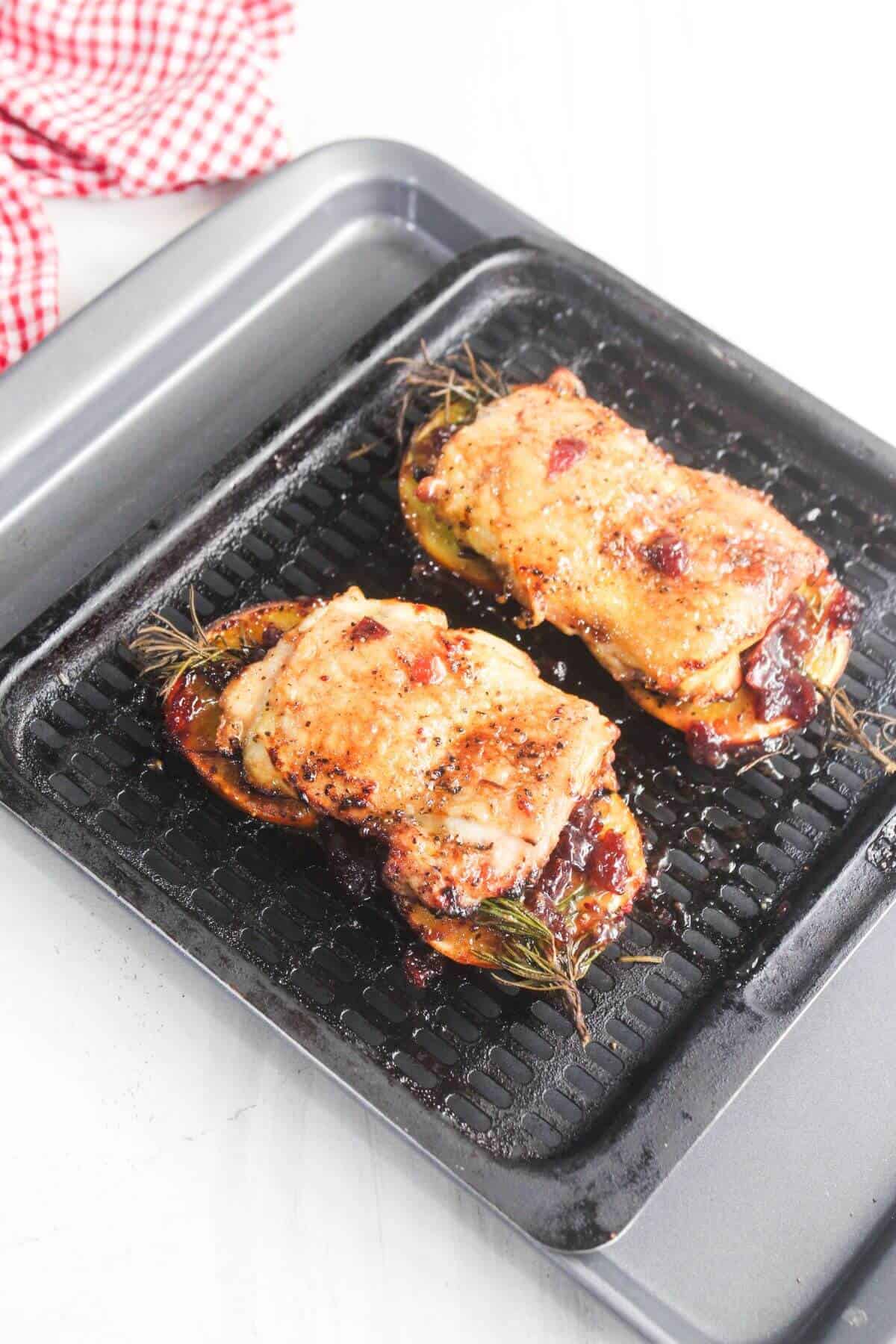 Two baked chicken thighs rest on a grill pan, each topped with herbs and glazed, with a checkered red cloth partially visible in the background.