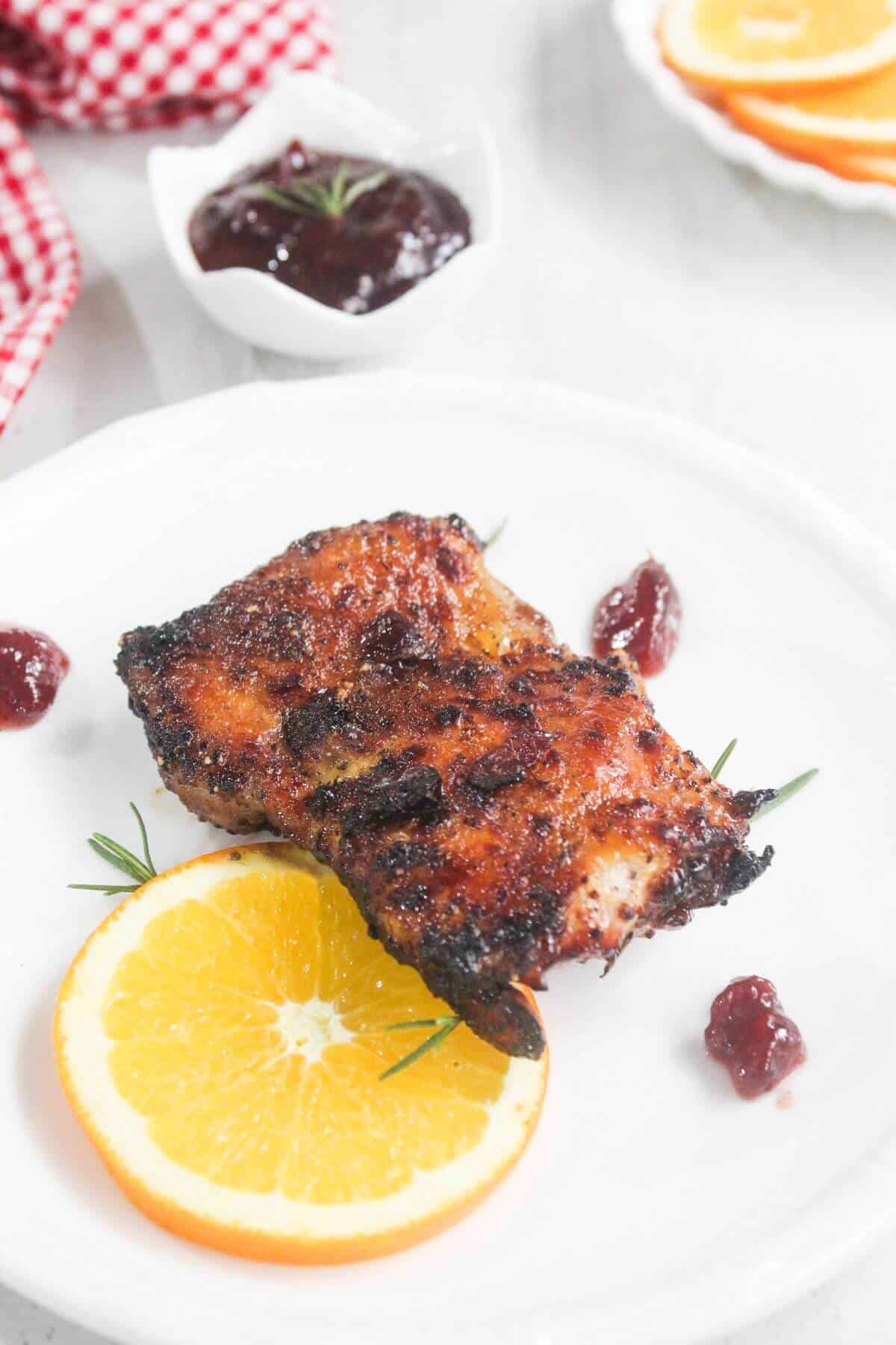 Grilled chicken thigh served on a white plate with an orange slice, garnished with rosemary and sauce dollops; extra sauce and orange slices in the background.