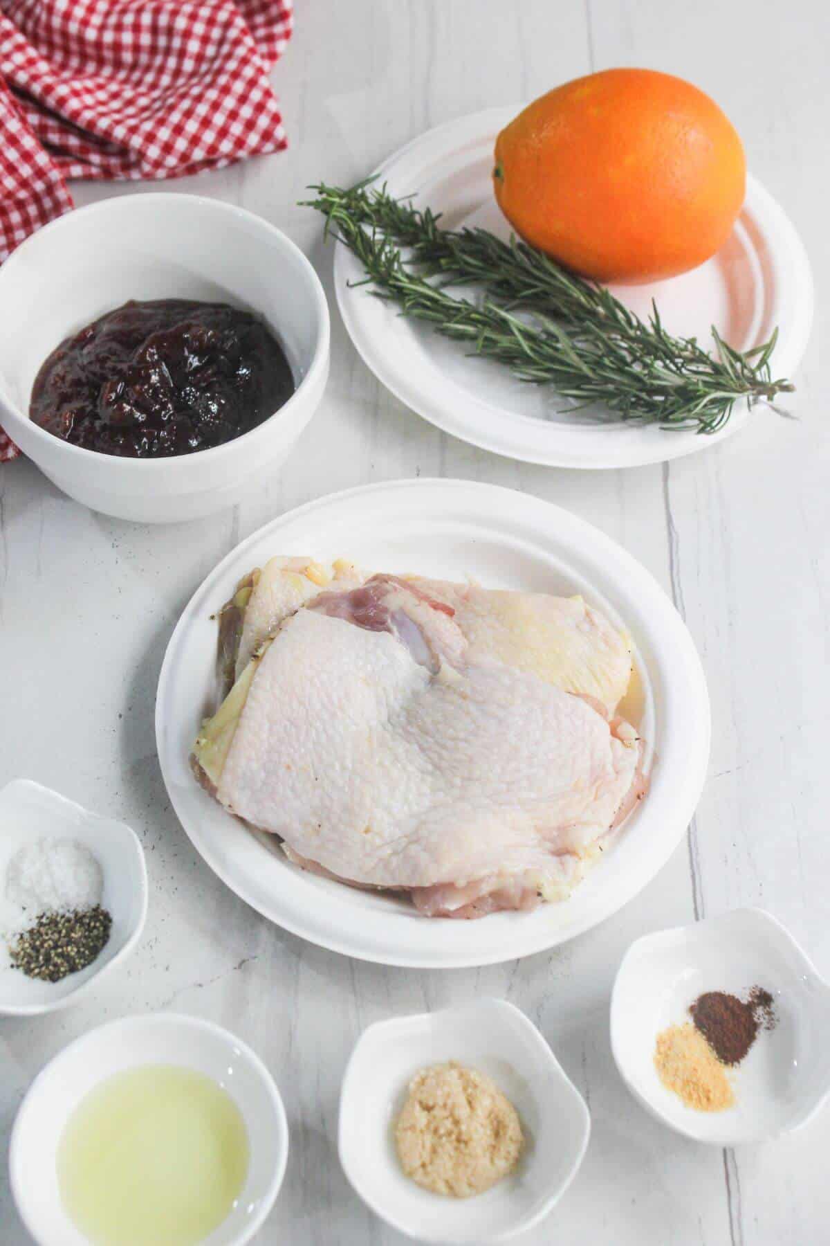 Raw chicken thighs on a plate surrounded by small bowls of spices, salt, pepper, lemon juice, brown sugar, jam, fresh rosemary, and an orange on a white surface.