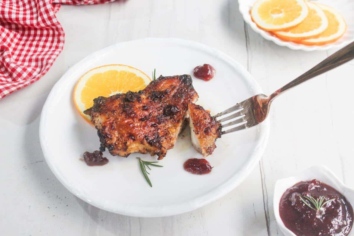 A plate with a grilled chicken thigh, orange slice, garnish, and dollops of sauce. A fork holds a piece of the chicken. Orange slices and sauce are in the background.