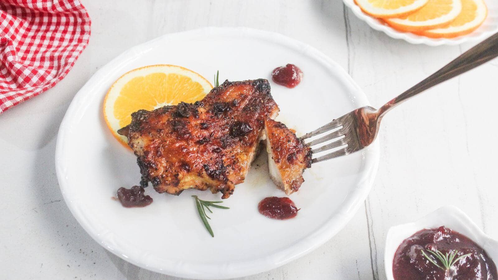 Grilled chicken breast with a slice of orange, small dollops of sauce, and a sprig of rosemary on a white plate. A fork is cutting into the chicken.
