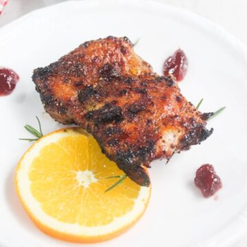 Two pieces of grilled meat served on a white plate with a slice of orange, small sprigs of rosemary, and three dollops of red sauce.