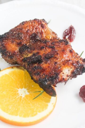 Two pieces of grilled meat served on a white plate with a slice of orange, small sprigs of rosemary, and three dollops of red sauce.