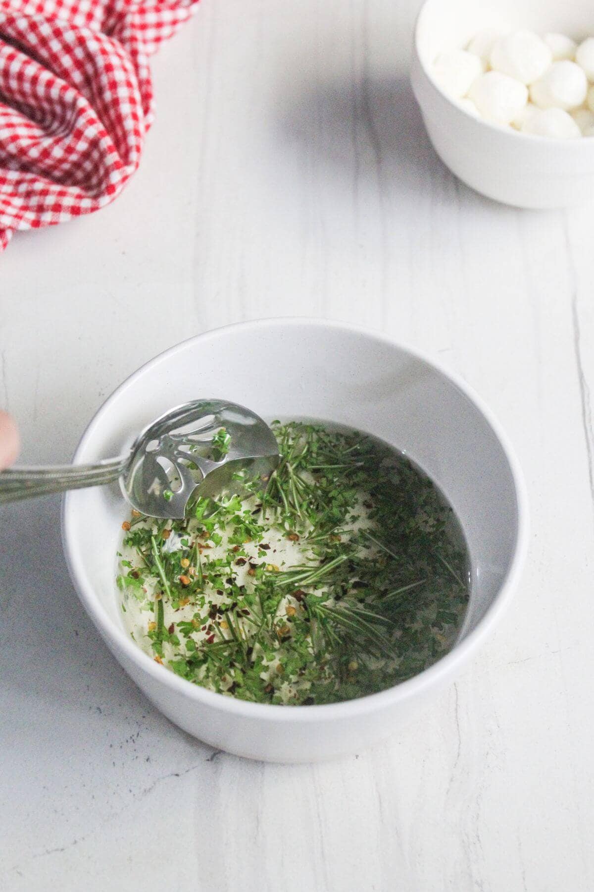 A white bowl with oil, chopped herbs, and spices being stirred with a spoon on a white surface; a red checkered cloth and another bowl are nearby.