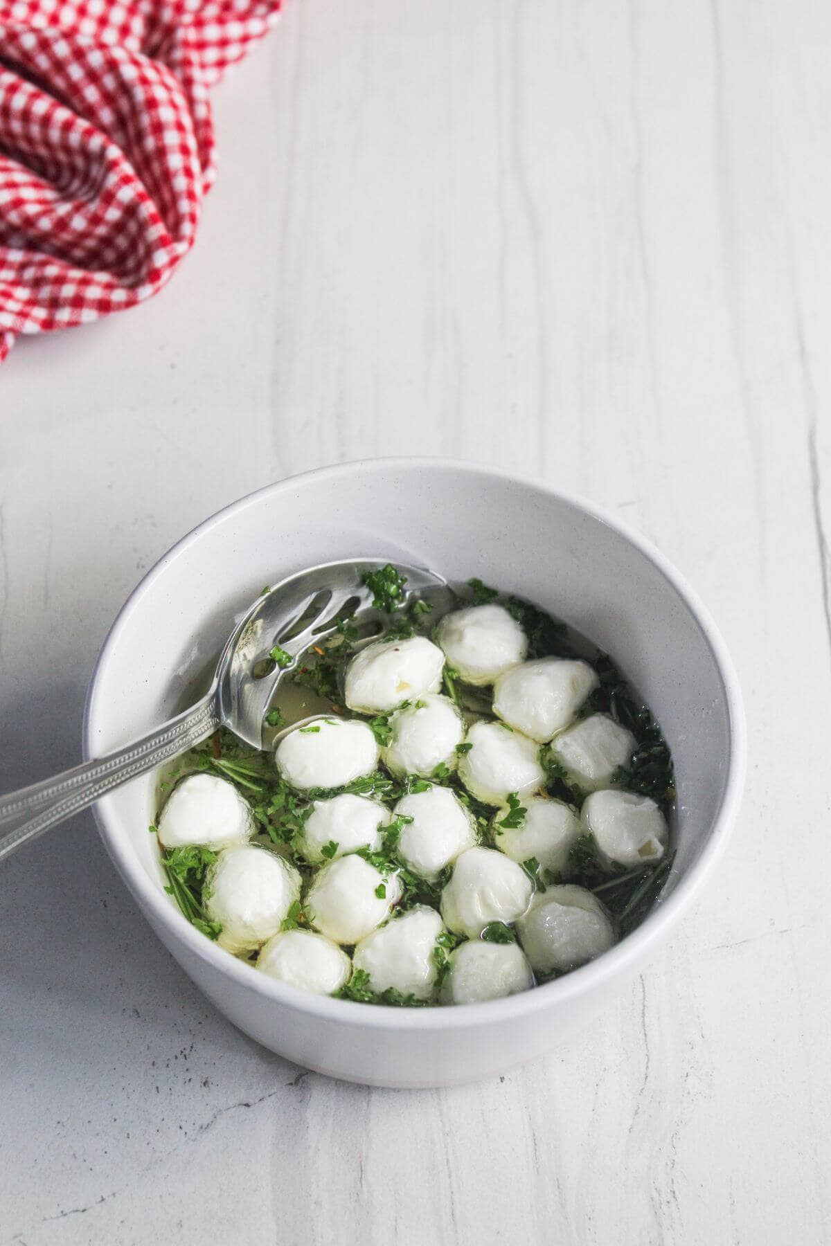 A white bowl filled with mozzarella balls in herb-infused oil with a slotted spoon, on a white surface. A red checkered cloth is partially visible in the background.