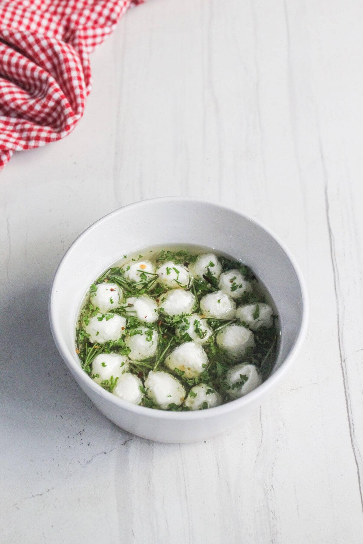A white bowl filled with clear soup containing small dumplings and herbs sits on a white surface next to a red checkered cloth.