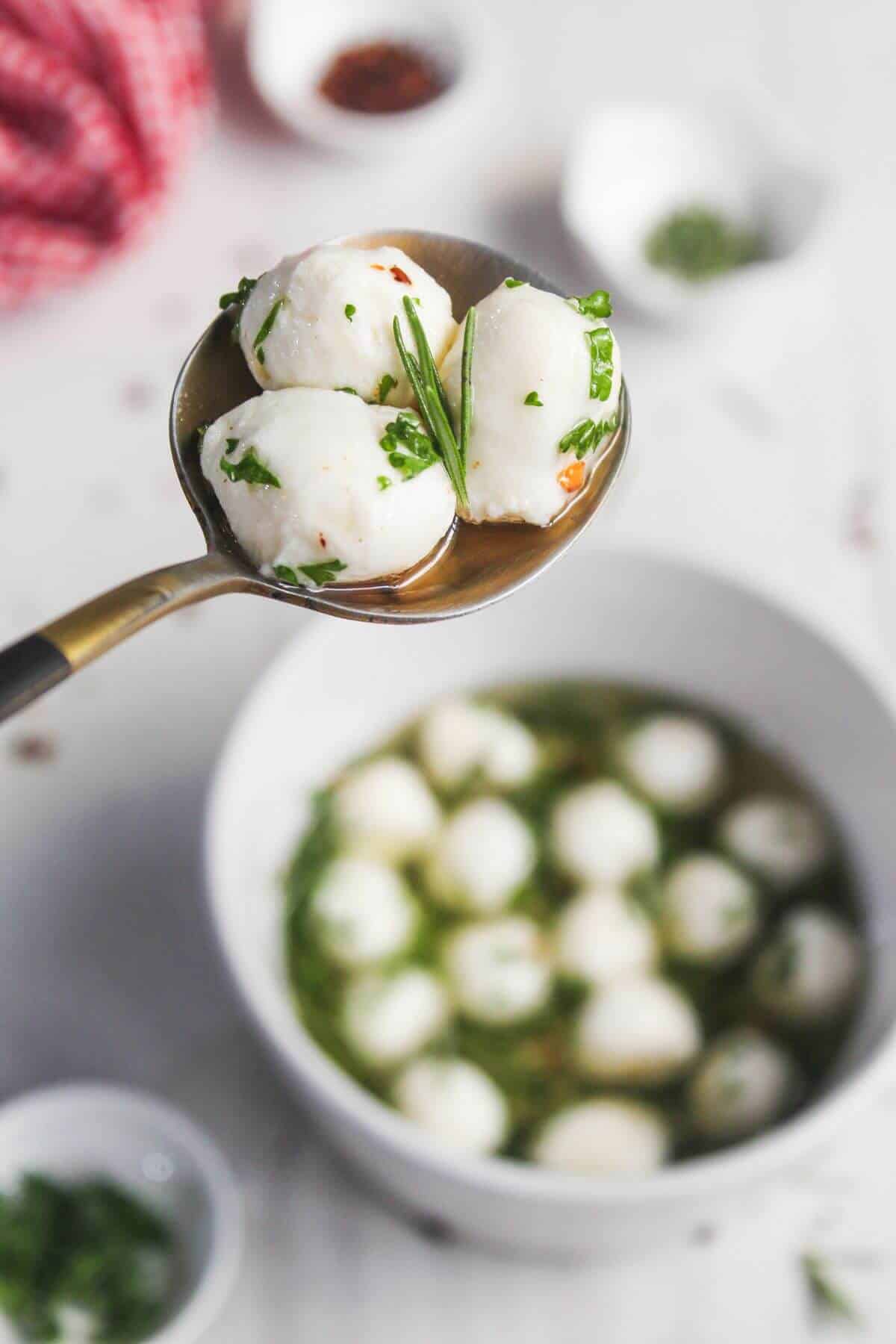 A spoon holds three mozzarella balls with herbs, above a bowl filled with more mozzarella balls and herbs in liquid.