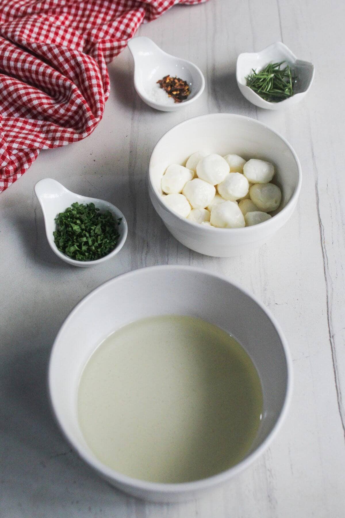 A bowl of creamy liquid, a bowl of mozzarella balls, and small dishes of chopped herbs, salt, and red pepper flakes on a white surface with a red checkered cloth.