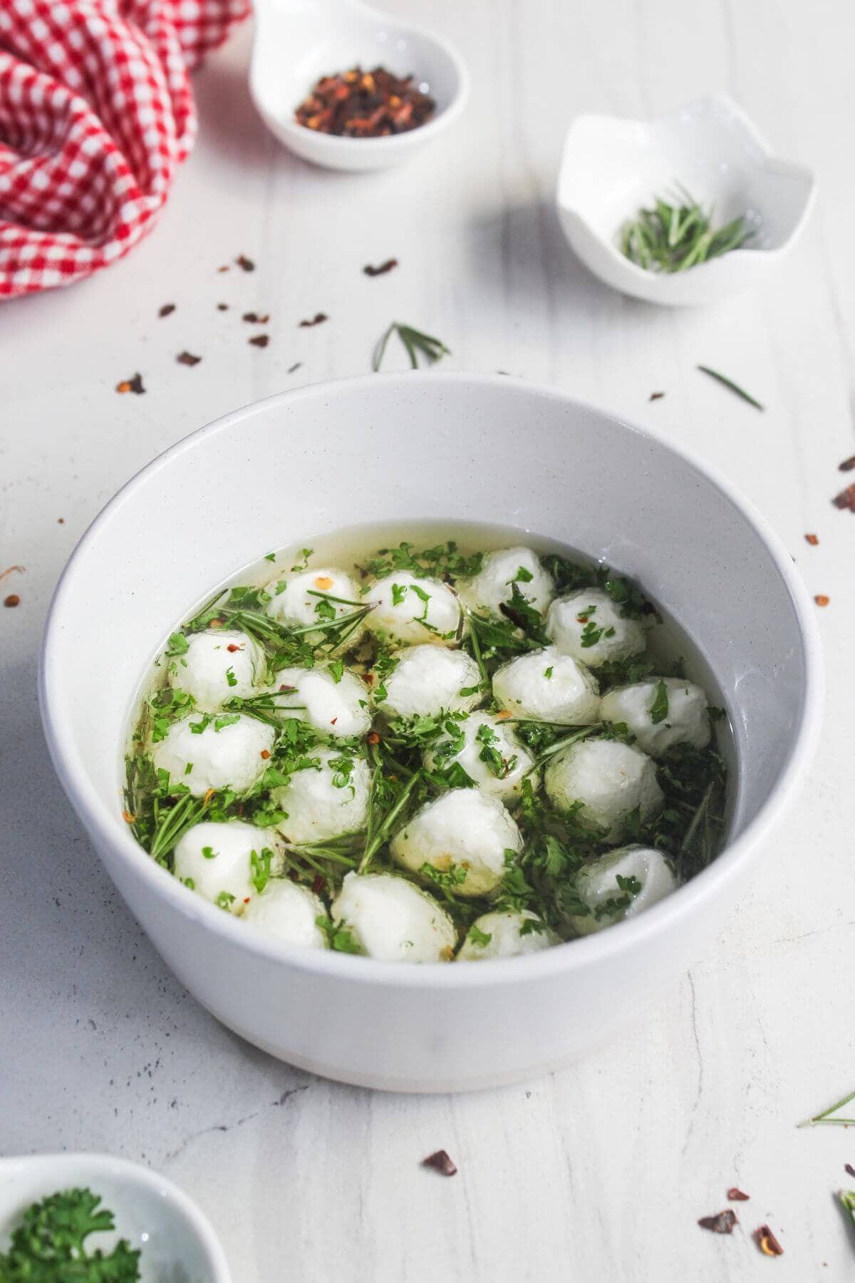 A white bowl filled with small mozzarella balls in herb-infused oil, garnished with fresh parsley and rosemary; small bowls of herbs and spices are in the background.