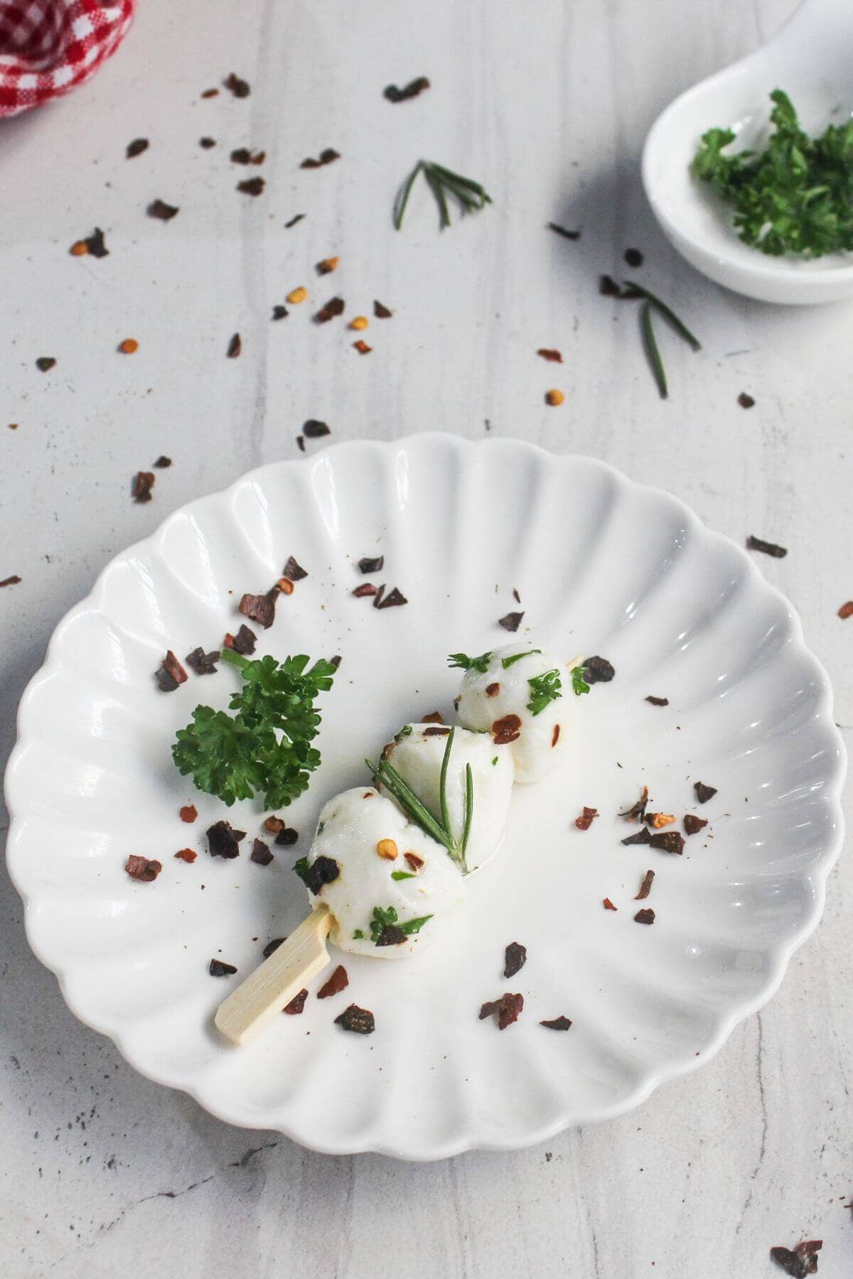 White scalloped plate with three mozzarella balls on a wooden stick, garnished with parsley, rosemary, and red pepper flakes. Some parsley and pepper flakes are scattered on the surface.