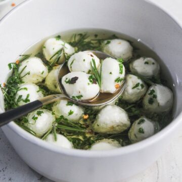 A bowl of mozzarella balls in olive oil with herbs, with a ladle scooping some out.