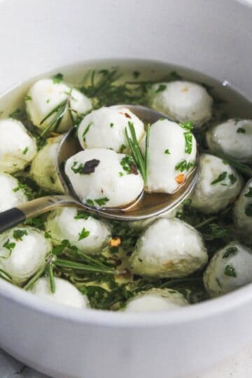 A bowl of mozzarella balls in olive oil with herbs, with a ladle scooping some out.