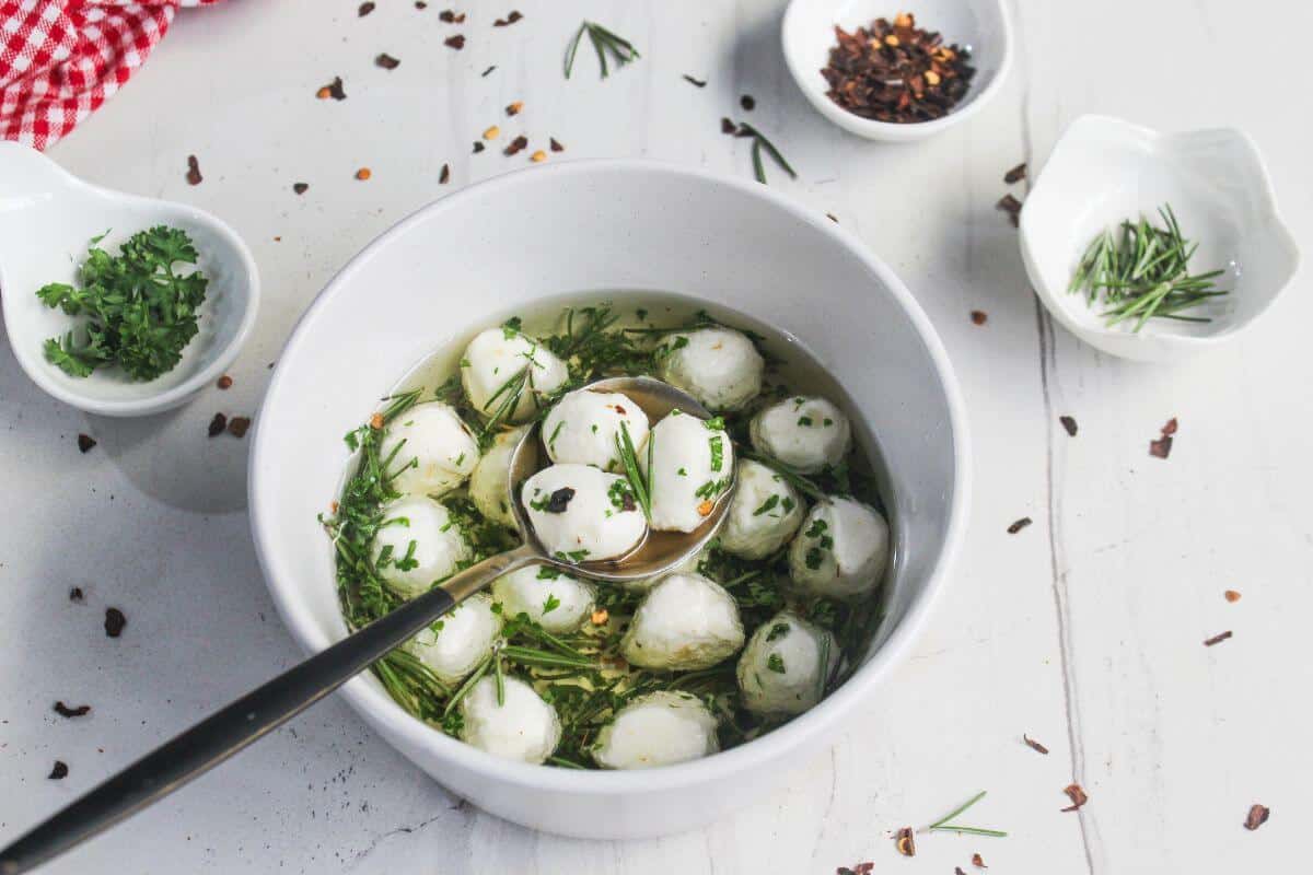A white bowl with round cheese balls in herb-infused oil, surrounded by small dishes of chopped herbs and chili flakes on a white surface.