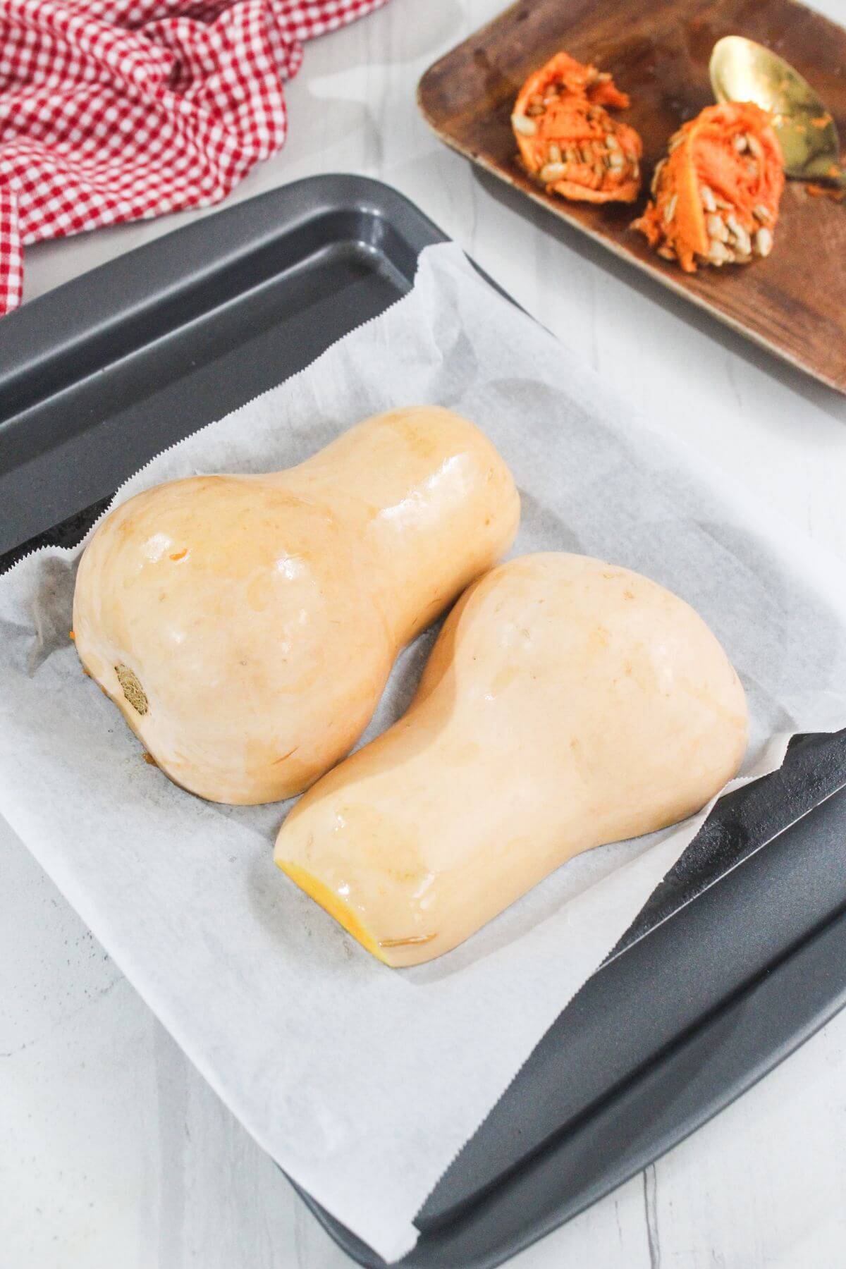 Two whole butternut squashes on a parchment-lined baking tray, with squash tops and a spoon on a brown plate in the background.