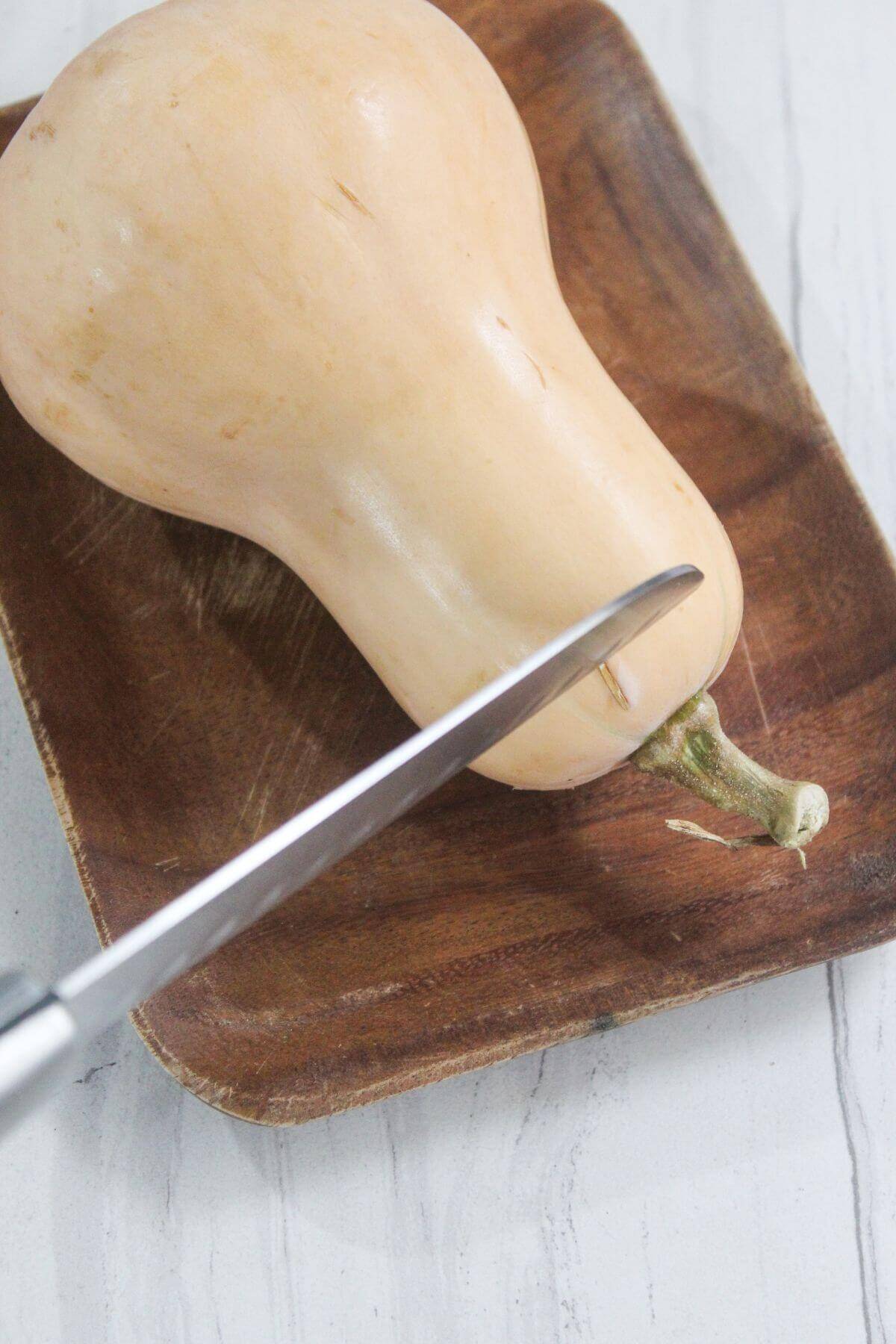 A knife is positioned to cut a whole butternut squash placed on a wooden cutting board.