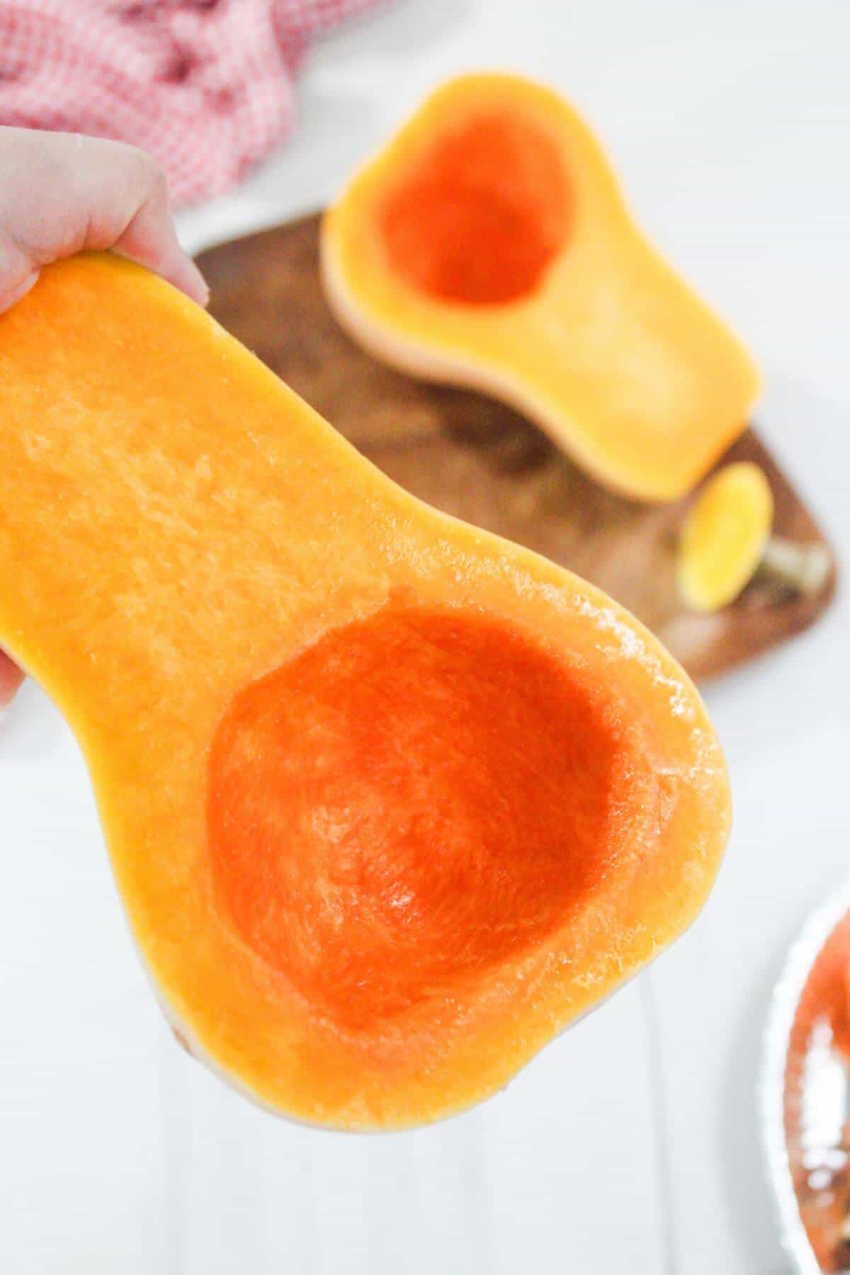 A hand holding a halved butternut squash with seeds removed; another half rests on a wooden cutting board in the background.