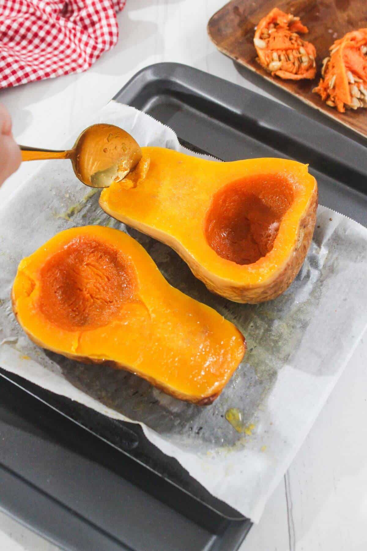 Two halves of roasted butternut squash on a baking tray lined with parchment paper; a spoon is scooping flesh from one half.