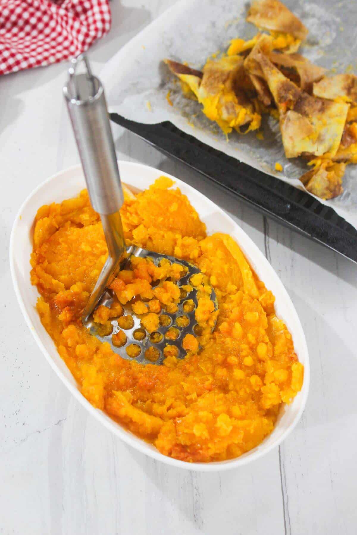 A metal masher pressing roasted pumpkin in a white oval dish, with pumpkin skins on a baking tray in the background.