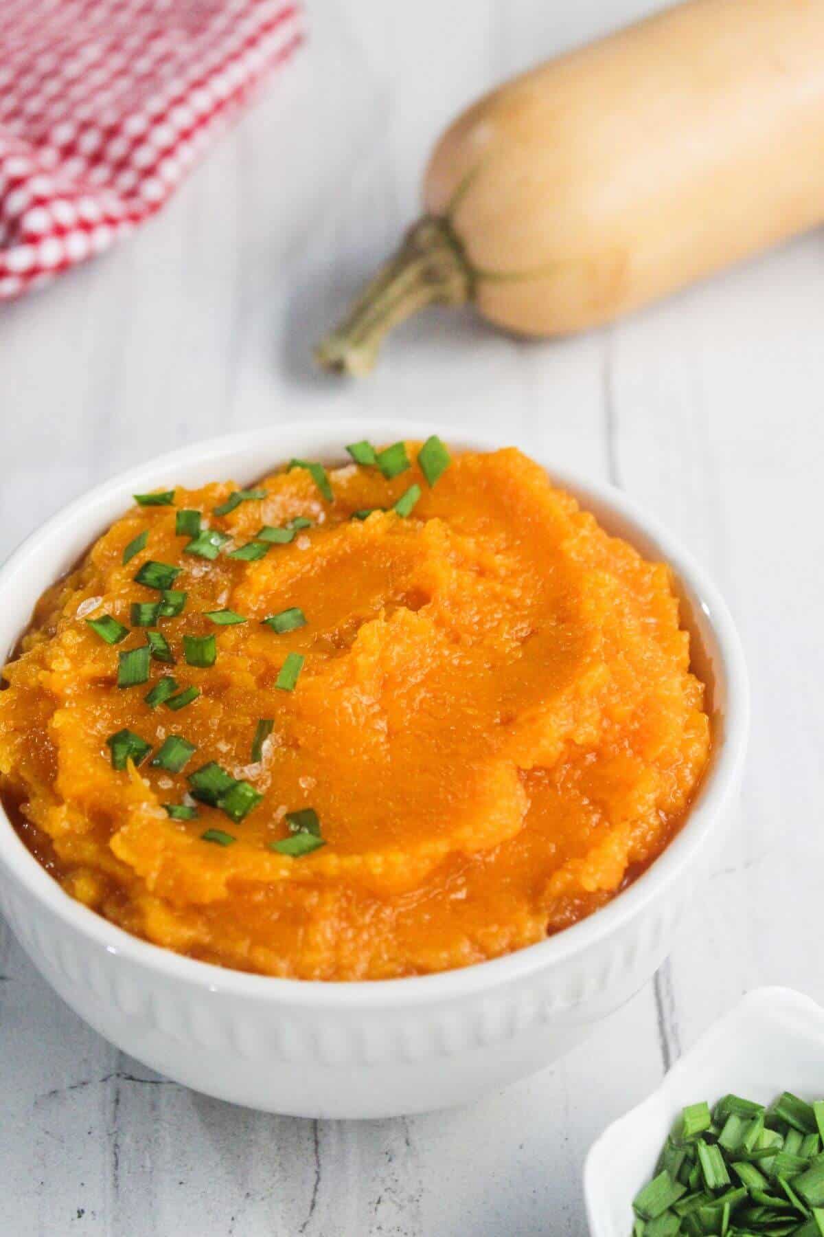 A white bowl filled with mashed butternut squash, garnished with chopped chives, sits on a white surface next to a whole butternut squash and a checkered cloth.