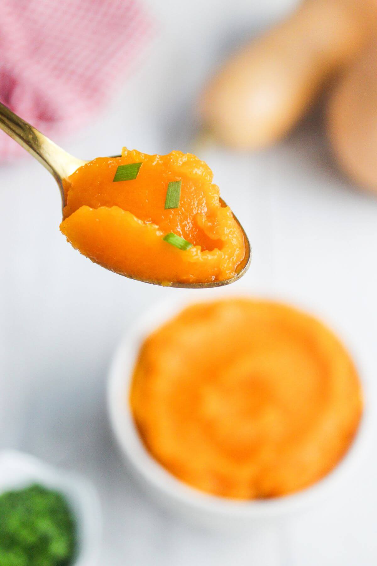 A spoonful of mashed butternut squash garnished with chopped green herbs, held above a bowl of more mashed squash.