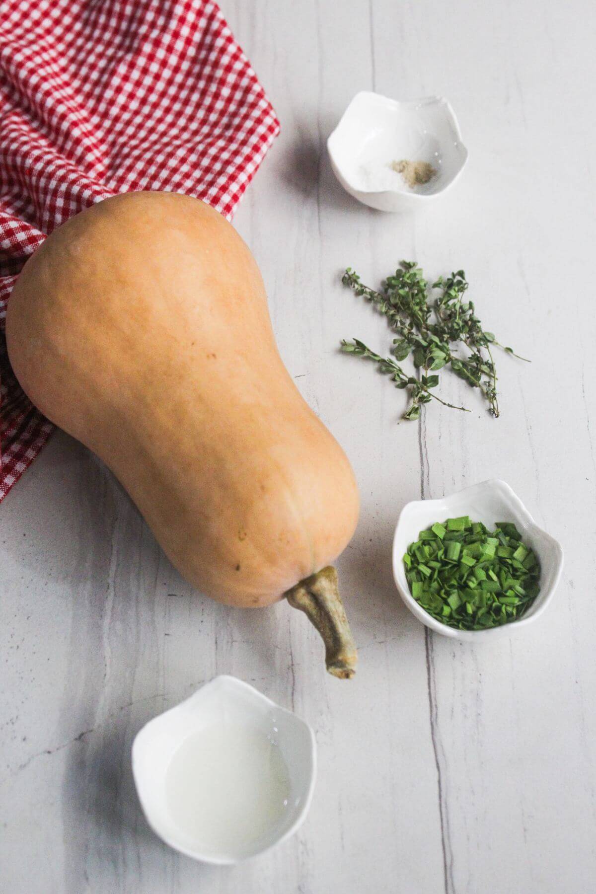 A whole butternut squash on a white surface with a red checkered cloth, fresh herbs, chopped scallions, a small bowl of liquid, and a small bowl of spices.