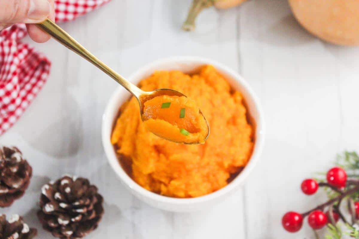 A bowl of bright orange mashed sweet potatoes is shown on a white table; a spoon lifts a portion topped with chopped chives. Pinecones and red berries are nearby.
