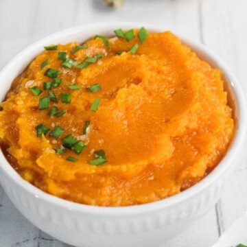 A white bowl filled with mashed sweet potatoes, garnished with chopped green herbs and coarse salt, on a white surface.