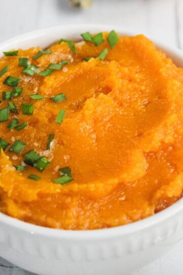 A white bowl filled with mashed sweet potatoes, garnished with chopped green herbs and coarse salt, on a white surface.