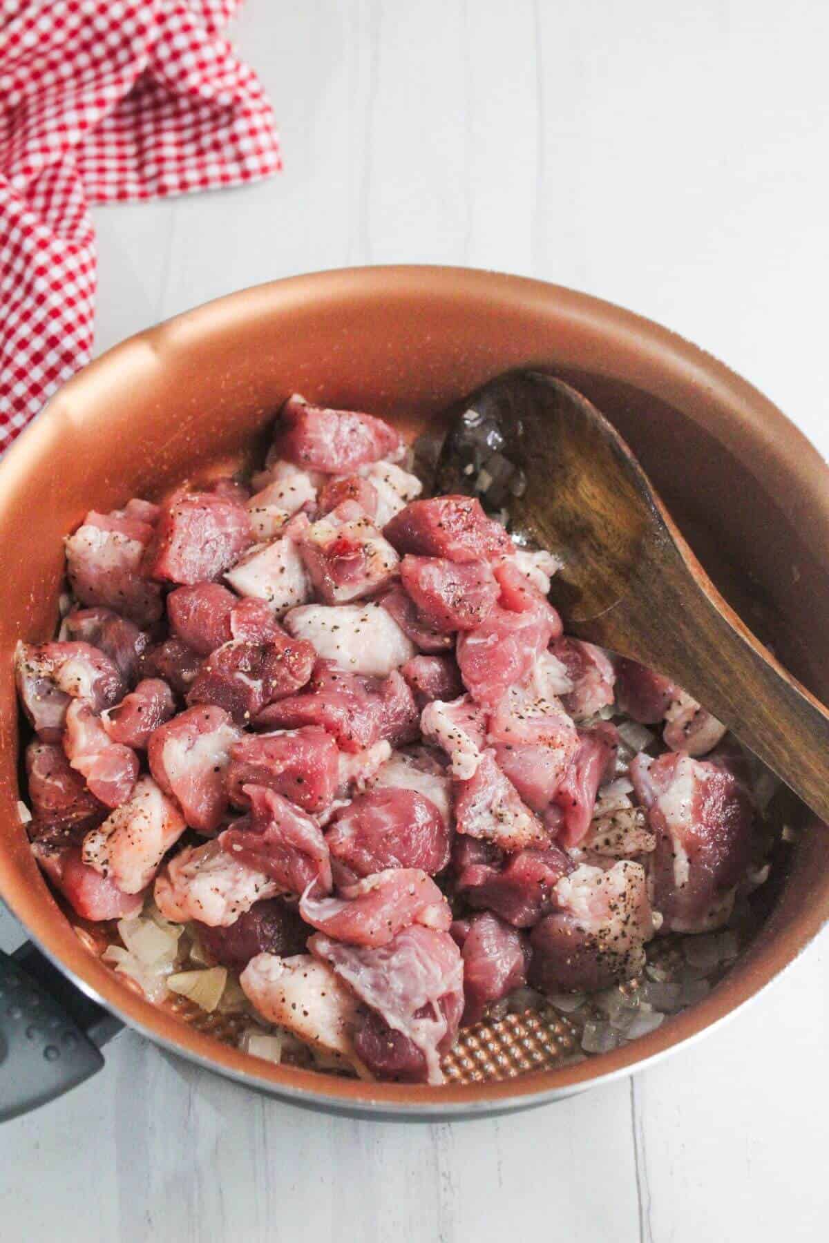 Raw, cubed meat with seasoning in a copper pan, sitting on top of chopped onions, with a wooden spoon resting on the side.