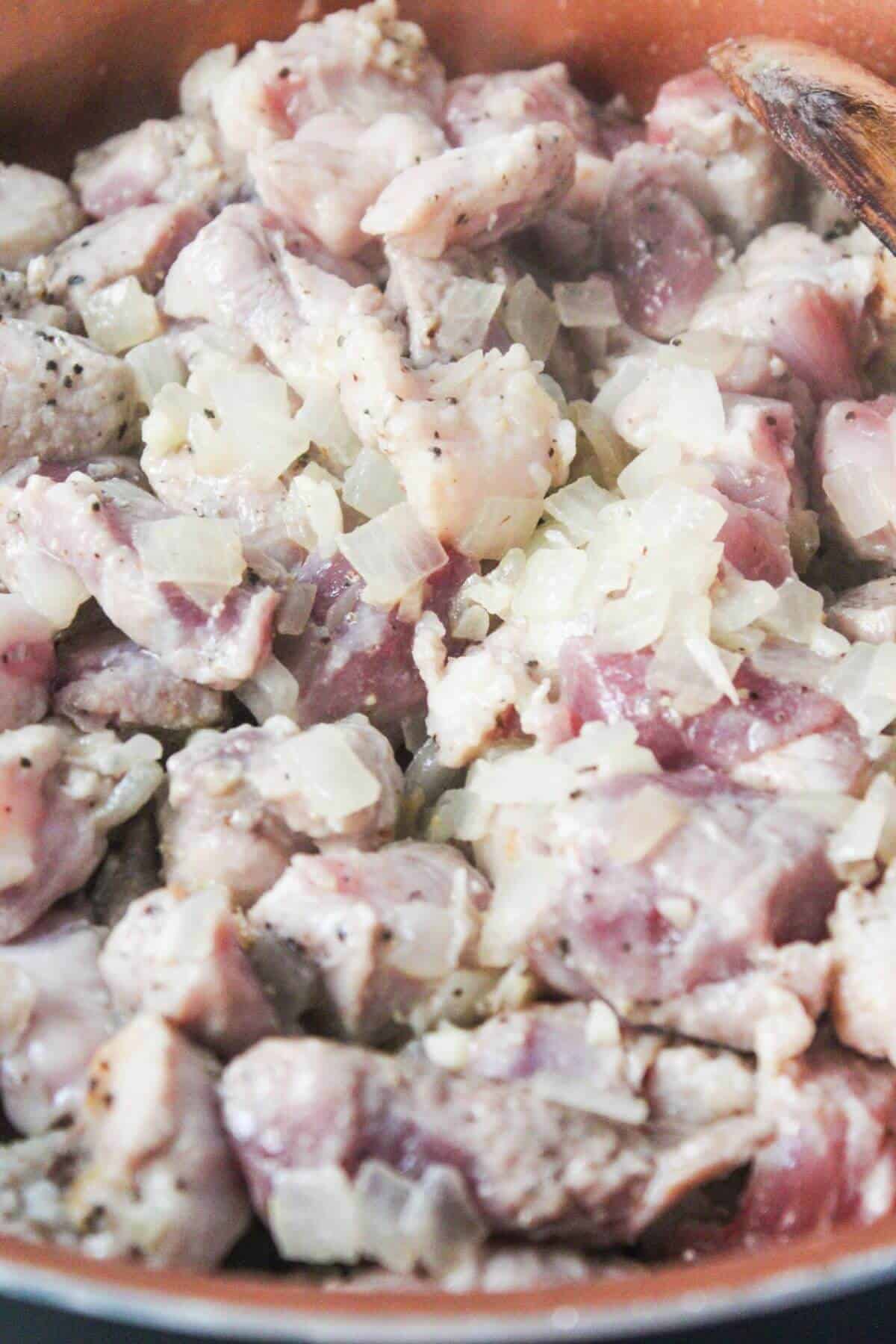 Diced raw pork and chopped onions being cooked in a pan with visible seasoning and a wooden spoon.