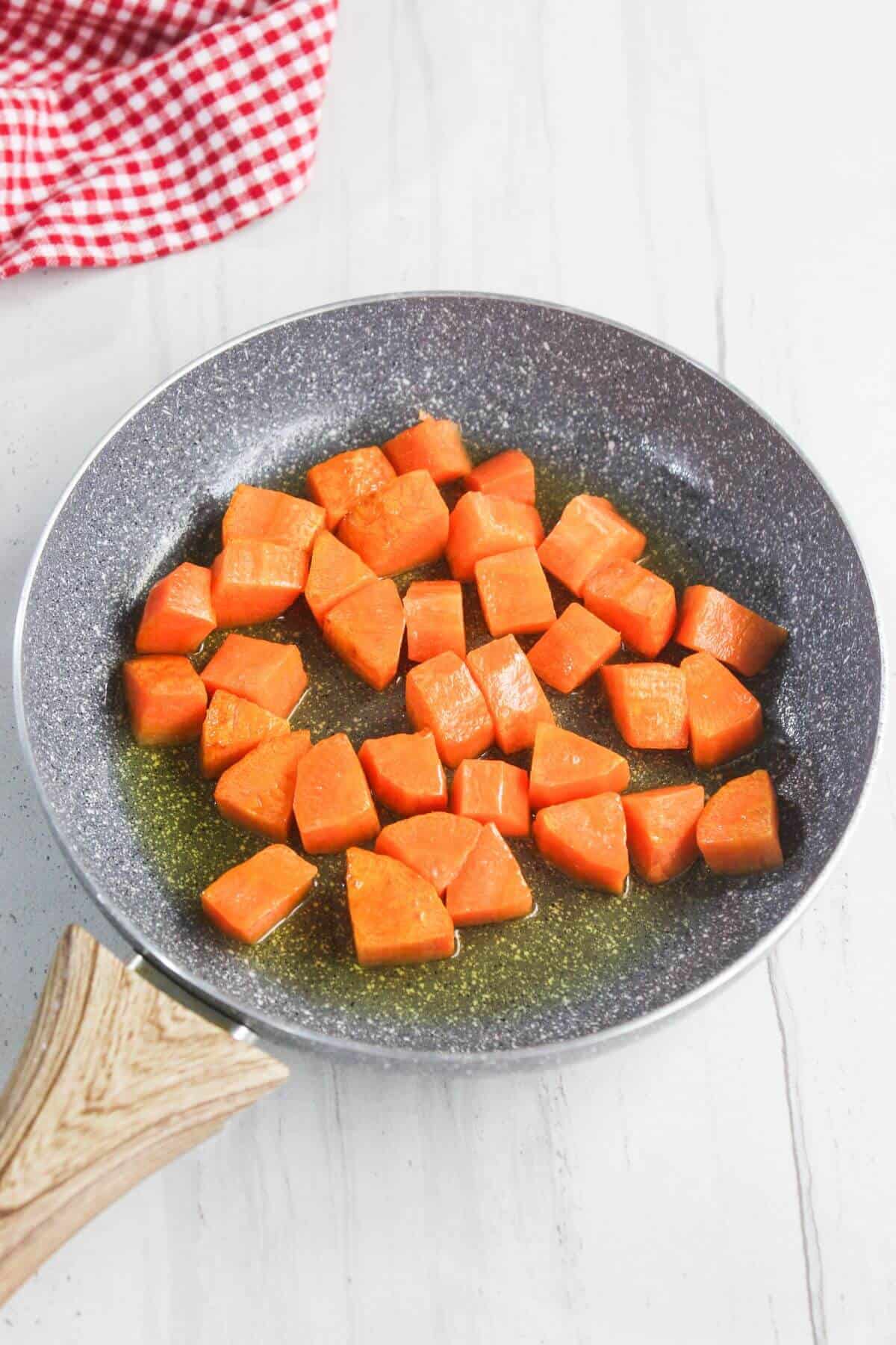Chopped carrots cooking in olive oil in a nonstick frying pan on a white surface, with a red and white checkered cloth in the background.