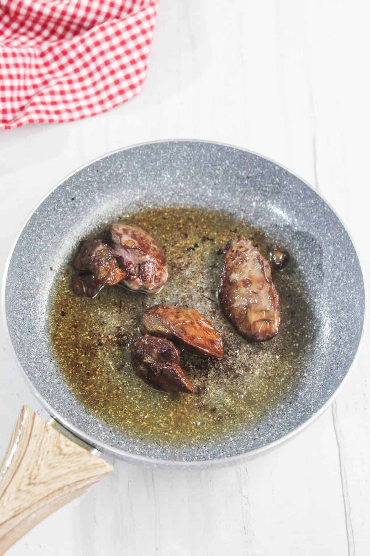 Four pieces of meat frying in oil in a grey nonstick pan on a white surface, with a red and white checkered cloth nearby.