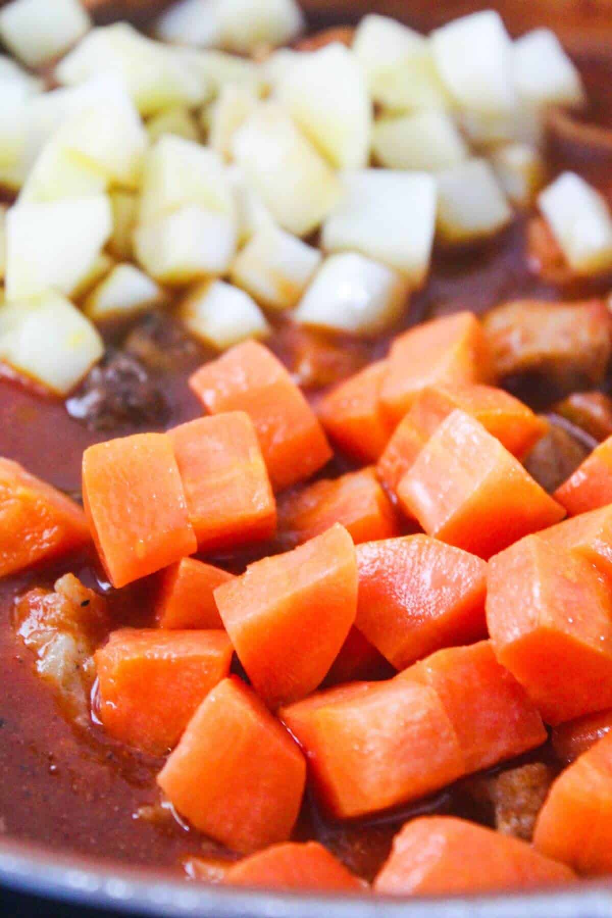 Close-up of diced carrots and potatoes being cooked in a red sauce.