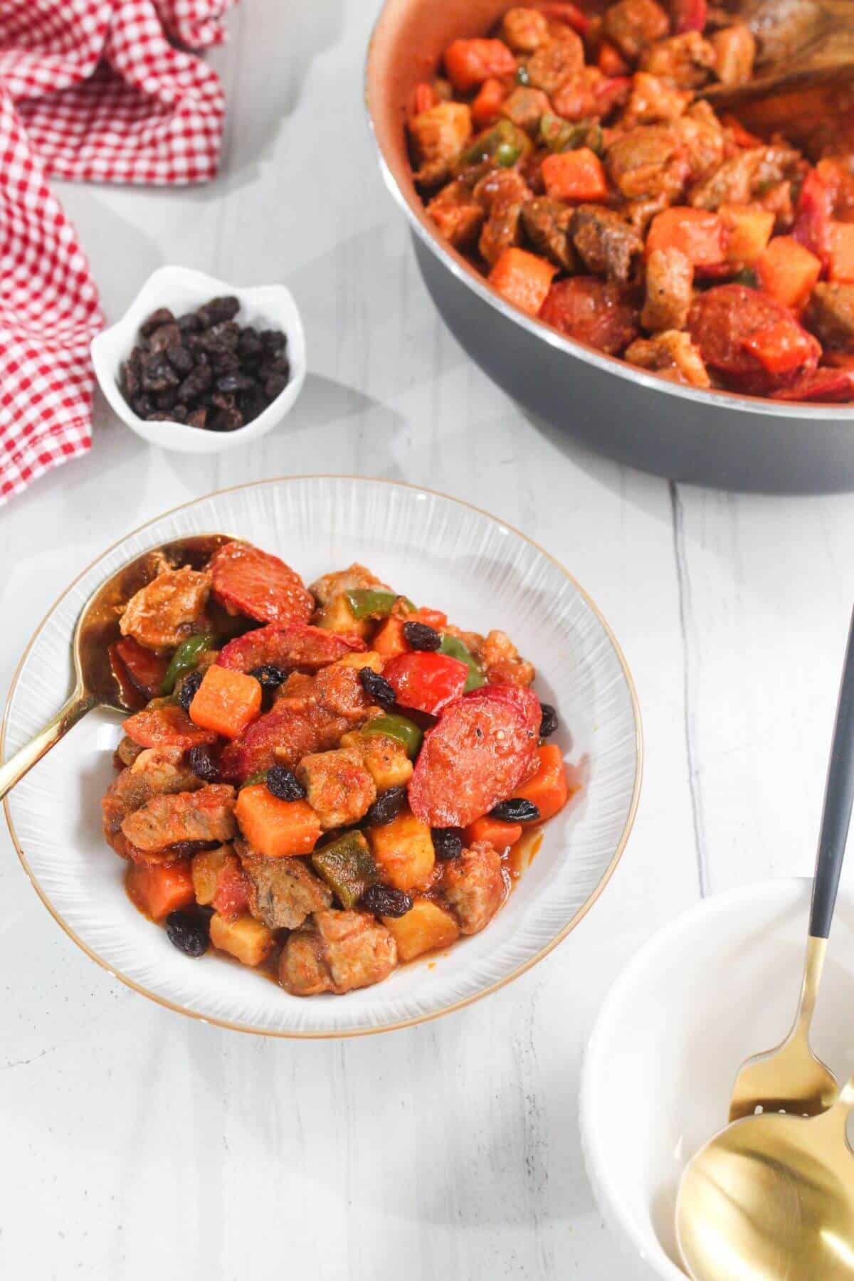 A bowl of pork menudo with vegetables and raisins sits on a white table next to a pan of the same dish, a small bowl of raisins, and a red checkered cloth.