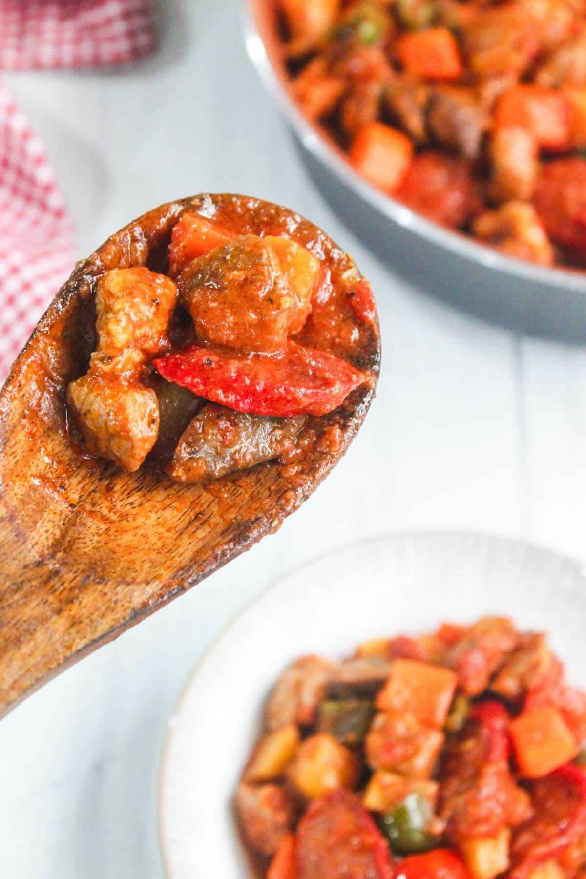 A wooden spoon holds a serving of chunky stew with meat, sausage, and vegetables, with bowls of the same stew in the background.