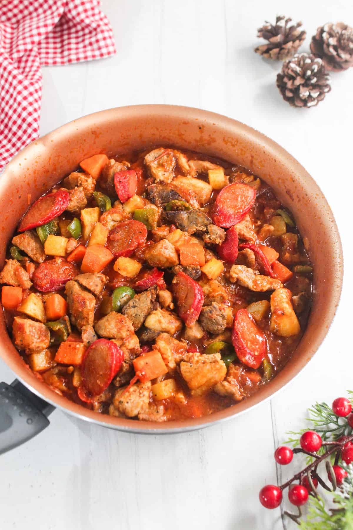 A pan of pork menudo with diced vegetables and sliced sausages in a red sauce, set on a white surface with pinecones, a red cloth, and festive greenery nearby showcases this classic Filipino pork menudo in a cozy holiday setting.