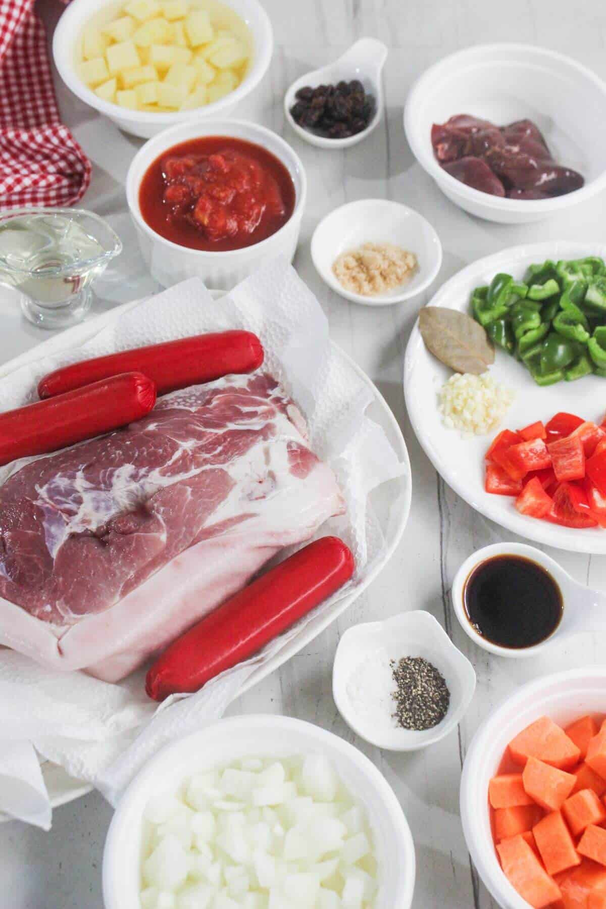 Assorted raw ingredients for a recipe, including pork, hotdogs, vegetables, seasonings, liver, tomato sauce, and small bowls of spices arranged on a white surface.