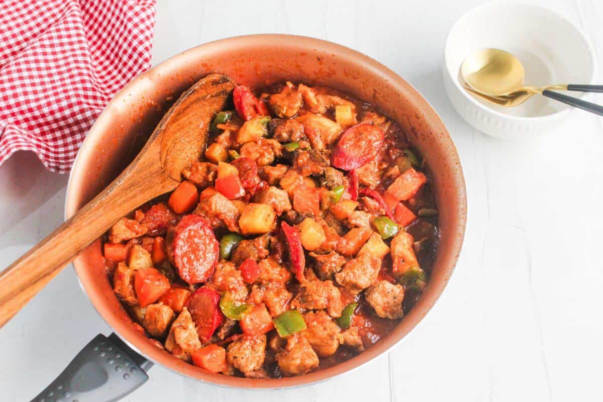 A pan filled with a cooked meat and vegetable stew, including chunks of sausage, carrots, potatoes, and bell peppers, with a wooden spoon resting inside. A bowl and utensils are nearby.