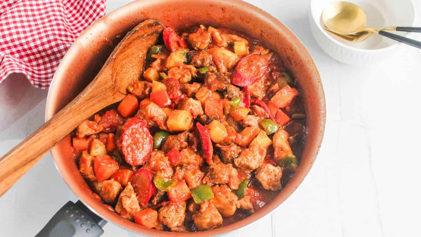 A pan of cooked pork menudo with chopped vegetables and sausage, mixed with sauce, sits on a white surface next to a spoon and empty bowl.