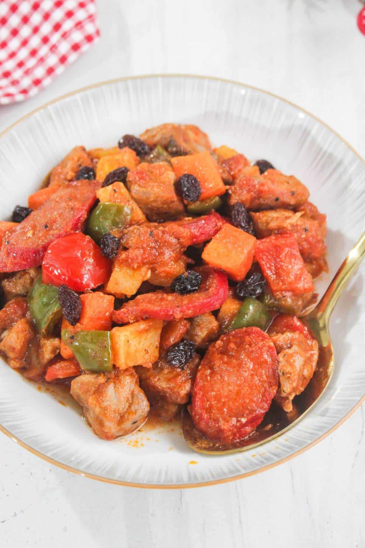 A plate of stew with chunks of meat, sausage, potatoes, carrots, bell peppers, and raisins in a red sauce, served with a spoon.