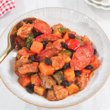A bowl of pork menudo with chunks of pork, potatoes, carrots, bell peppers, sliced sausages, and raisins in a tomato-based sauce, served with a spoon.