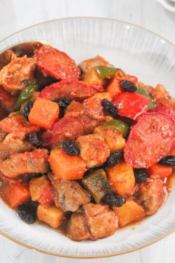 A bowl of pork menudo with chunks of pork, potatoes, carrots, bell peppers, sliced sausages, and raisins in a tomato-based sauce, served with a spoon.