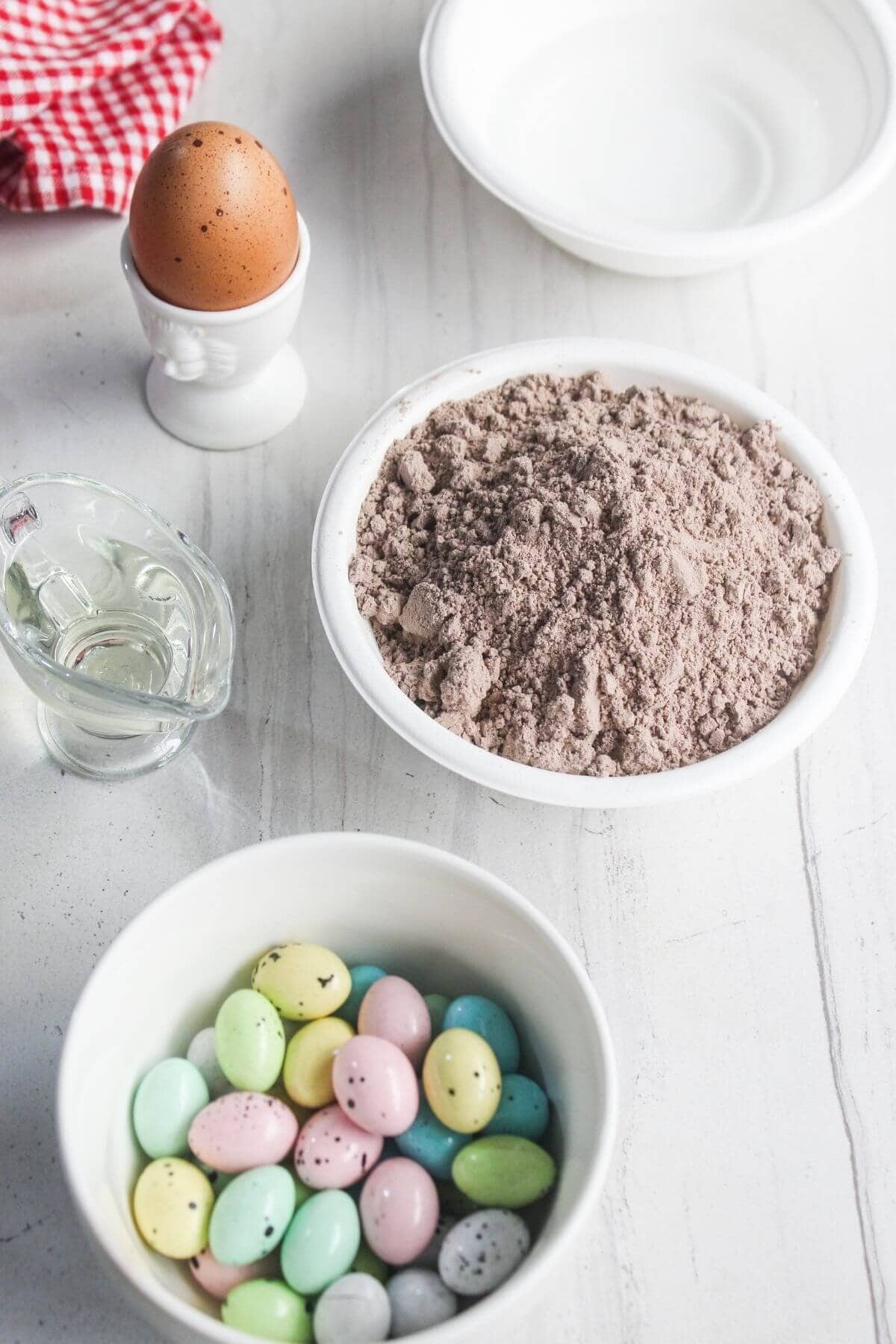 A bowl of chocolate cake mix, a bowl of pastel candy eggs, a whole egg in an egg cup, a small cup of oil, and an empty bowl on a white surface.