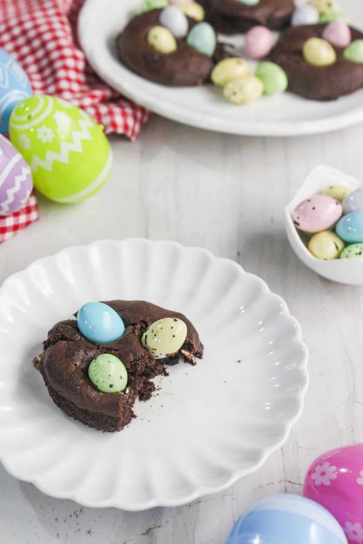 A chocolate cookie topped with pastel candy eggs is partially eaten on a white plate, surrounded by decorated Easter eggs and a bowl of candies.